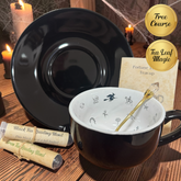 A black ceramic tea cup with a white interior, shown with a matching black saucer, reading blends, a gold spoon, and promotional badges reading 'Free Course' and 'Tea Leaf Magic' on a rustic wooden table with candles and spiderweb decor.