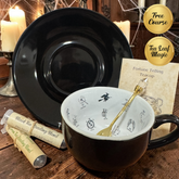 A witch-themed tea cup set displayed on a rustic wooden table, featuring a black teacup with a white interior decorated with black illustrations, a matching black saucer, two tubes of tea blending powder, and a fortune telling card. Two gold circular badges on the right read 'Free Course' and 'Tea Leaf Magic'.
