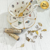 A styled flat-lay of a white porcelain teacup and saucer with gold trim and illustrated motifs, a gold spoon inside the cup, several silver and gold tea-themed charms, a rolled parchment label reading 'Black Tea Reading Blend,' and a gold circular badge that says 'Free Course' on a white wood-textured background.