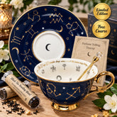 A luxury tea teacup and saucer set with a celestial theme, featuring a deep navy blue glaze decorated with gold constellations, stars, and crescent moons, along with a gold spoon. The set is styled on a wooden table with tea tubes, white flowers, and promotional badges.