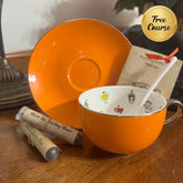 An orange ceramic teacup and saucer set on a wooden table, with a white spoon inside the cup, two wrapped tea packets, and a gold 'Free Course' badge in the top right corner.