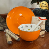 An orange ceramic teacup and saucer set with a white interior decorated with small colorful illustrations, a white spoon, and a small gift card. Two rolled tea sachets lie on a wooden surface beside the cup, with decorative objects and a plant in the background.