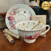 A white porcelain teacup and saucer set with light blue stripes, pink floral prints, and small black illustrations, displayed on a wooden table with tea bags and a gold 'Free Course' badge.