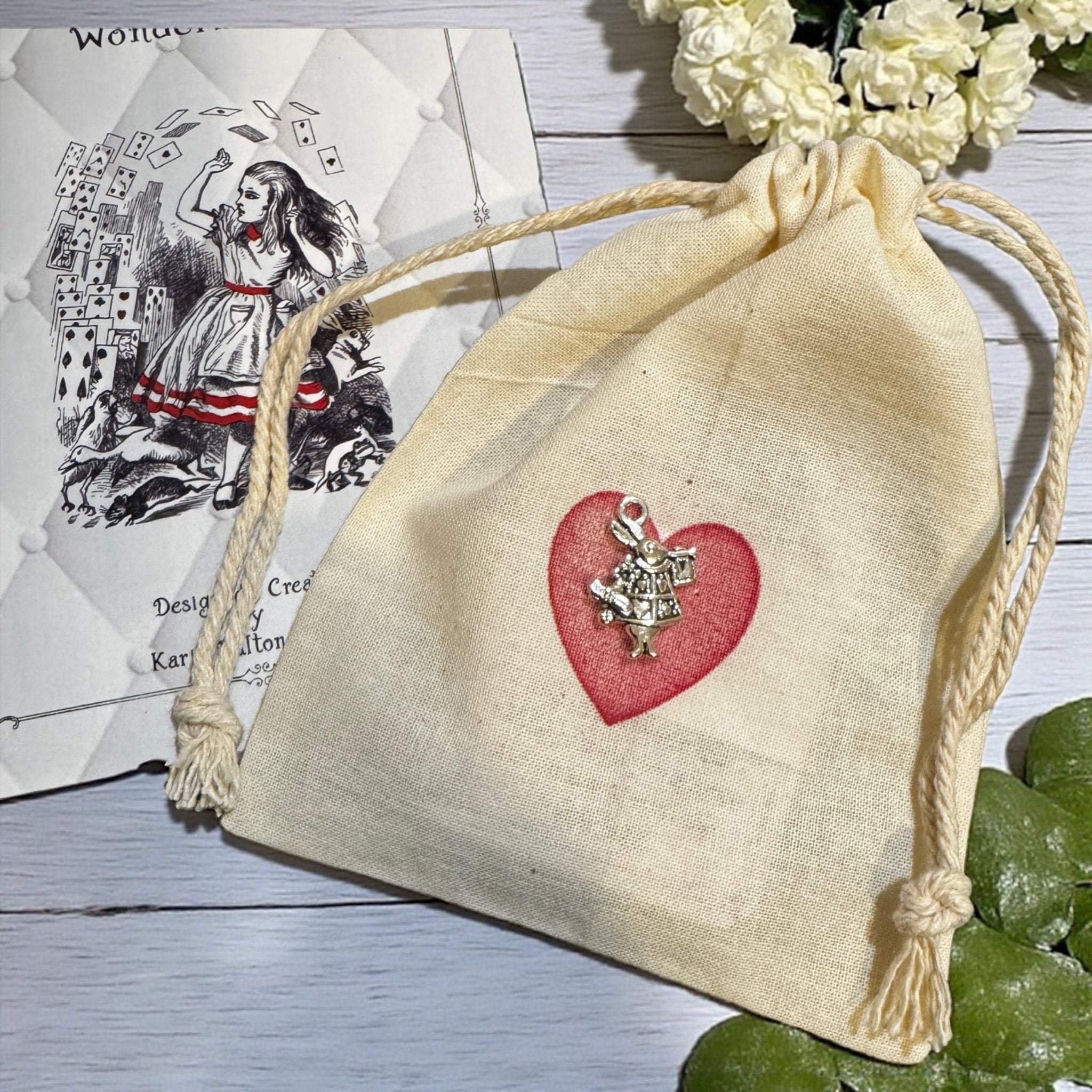Beige drawstring bag with a red heart and charm on a wooden surface with a card and flowers in the background.