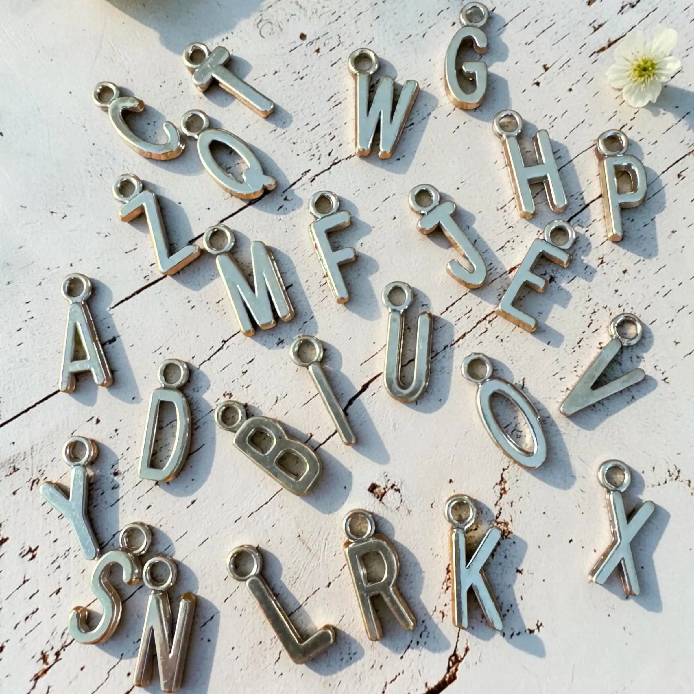 Alphabet letter charms in a distressed silver finish scattered on a white wooden surface with a small white flower in the corner.