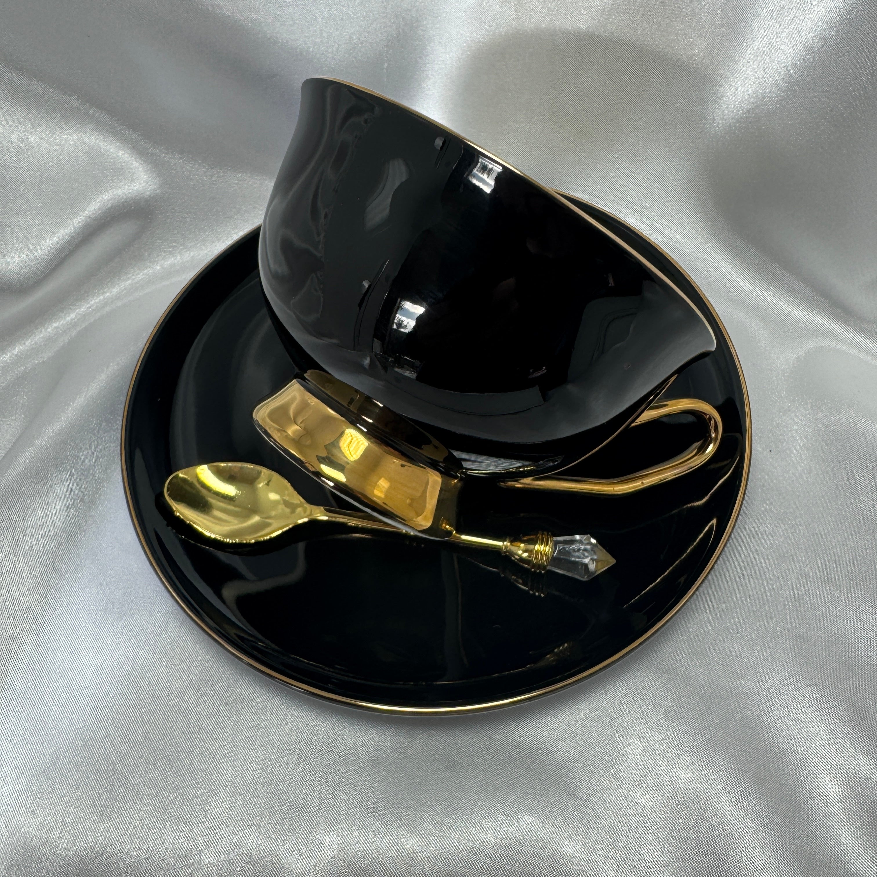 Side view of a glossy black teacup with a gold handle and base, sitting on a matching black saucer with a gold rim, alongside a gold spoon.