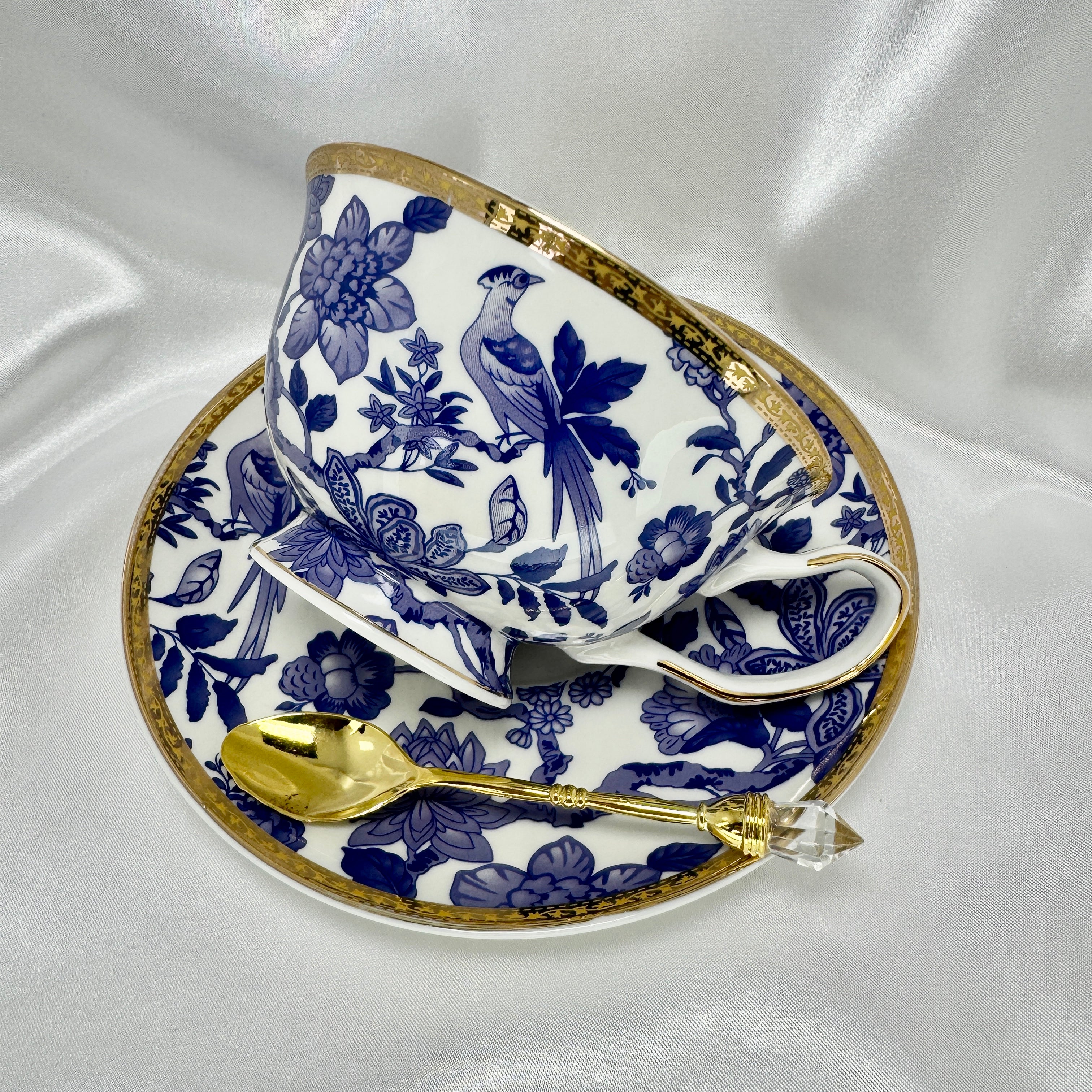 Bone china teacup and saucer in blue and white floral and bird pattern with gold accents, set against white satin.