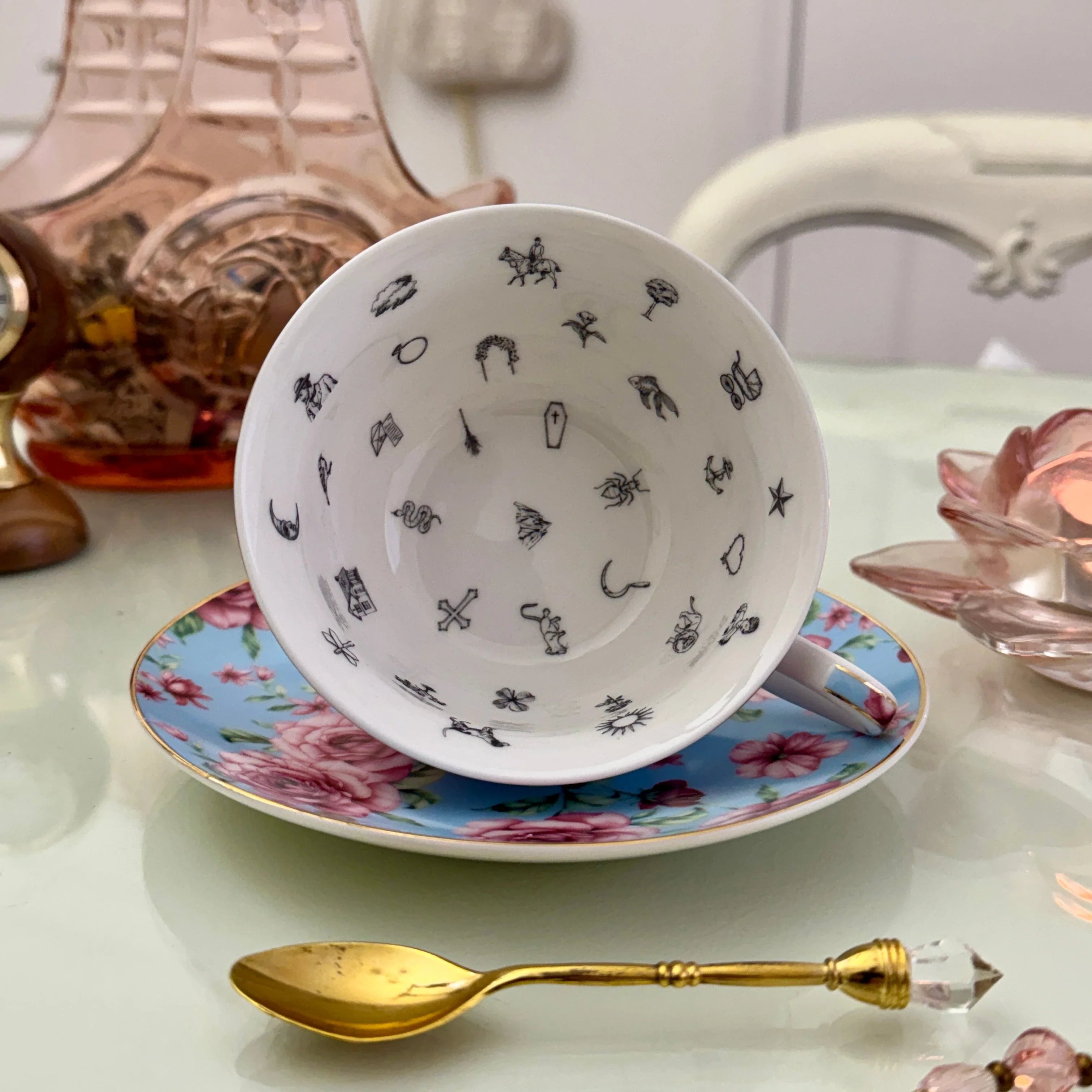 Inside view of blue pink rose floral tasseography teacup face-up showing all 32 printed tea leaf reading divination symbols, resting on matching saucer with gold crystal teaspoon