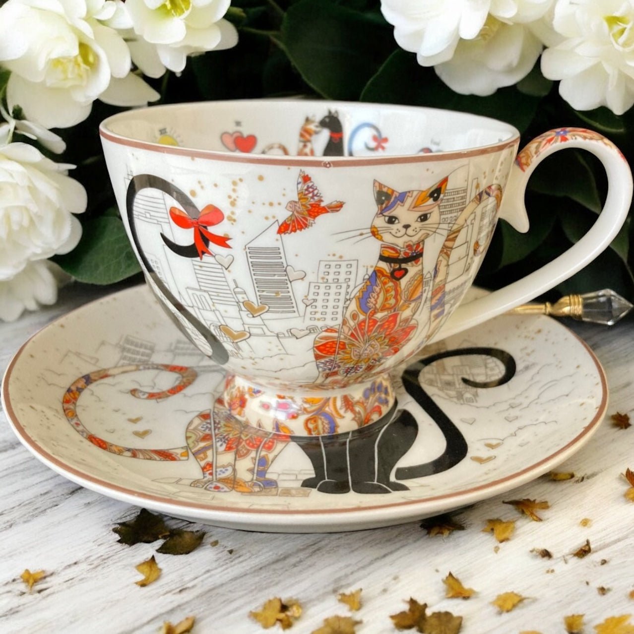 Decorative teacup and saucer with cat design on a wooden surface with flowers in the background