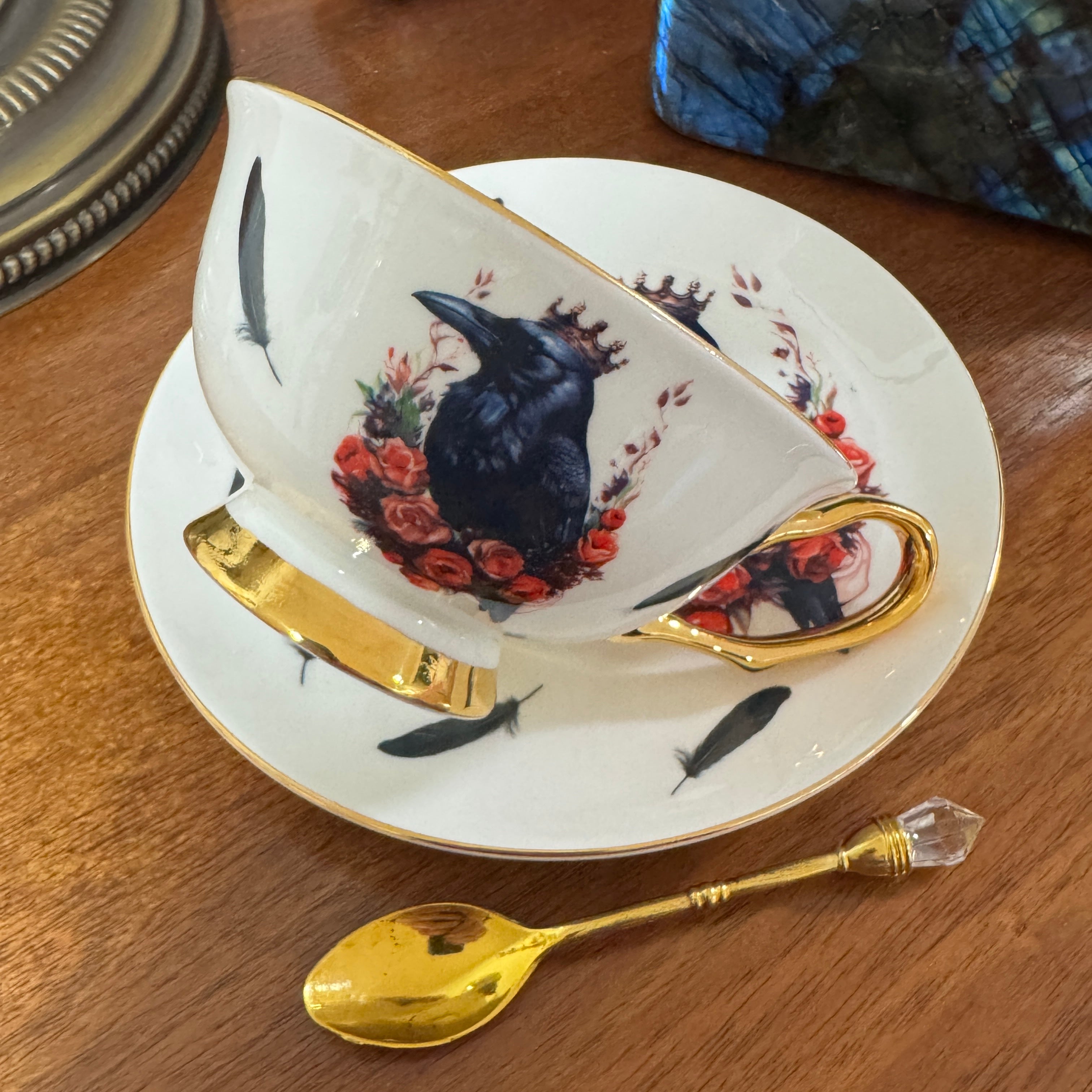 Teacup with raven and floral design on a wooden table with a gold spoon.