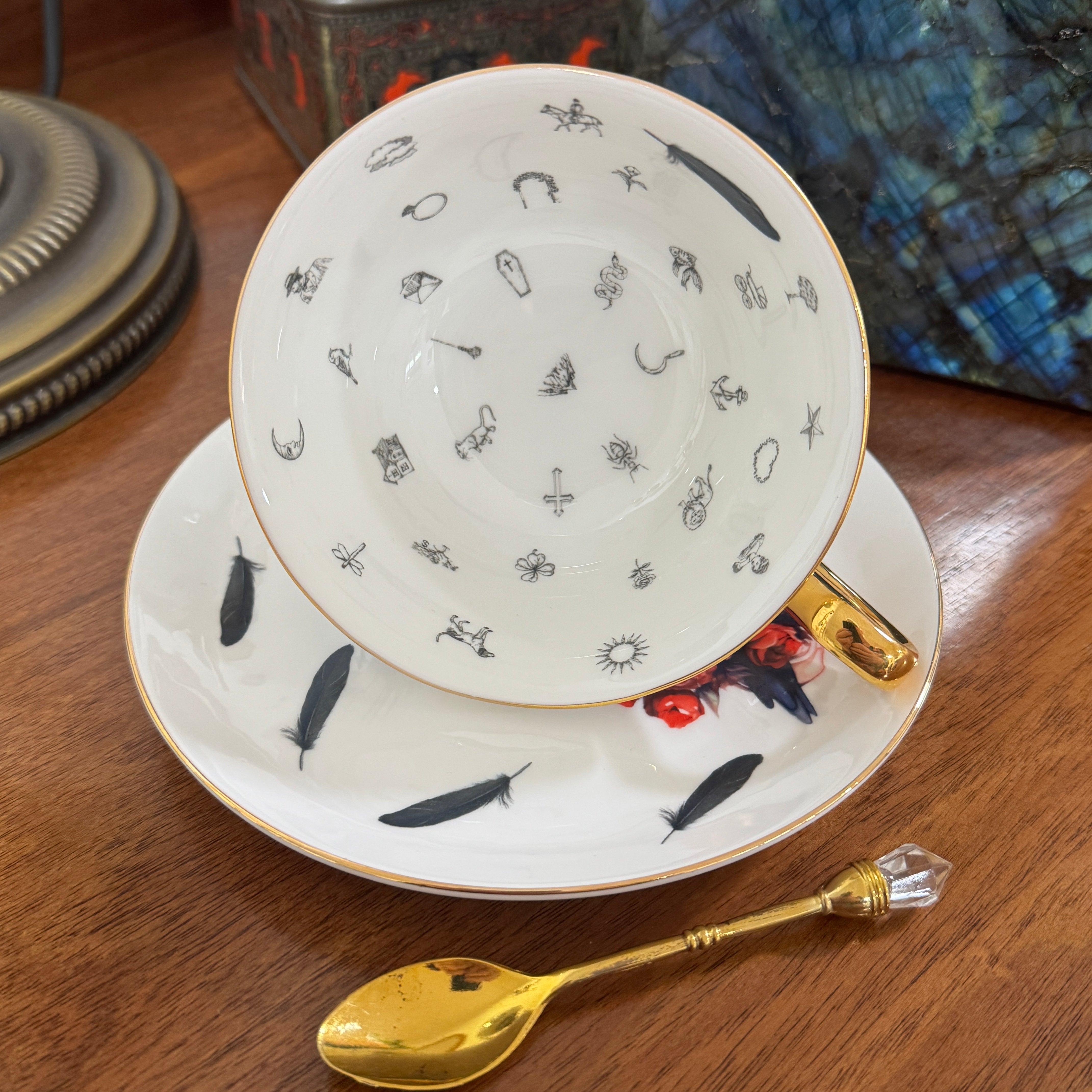 Teacup and saucer with feather pattern on a wooden surface, accompanied by a gold spoon.