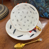 Teacup and saucer with feather pattern on a wooden surface, accompanied by a gold spoon.