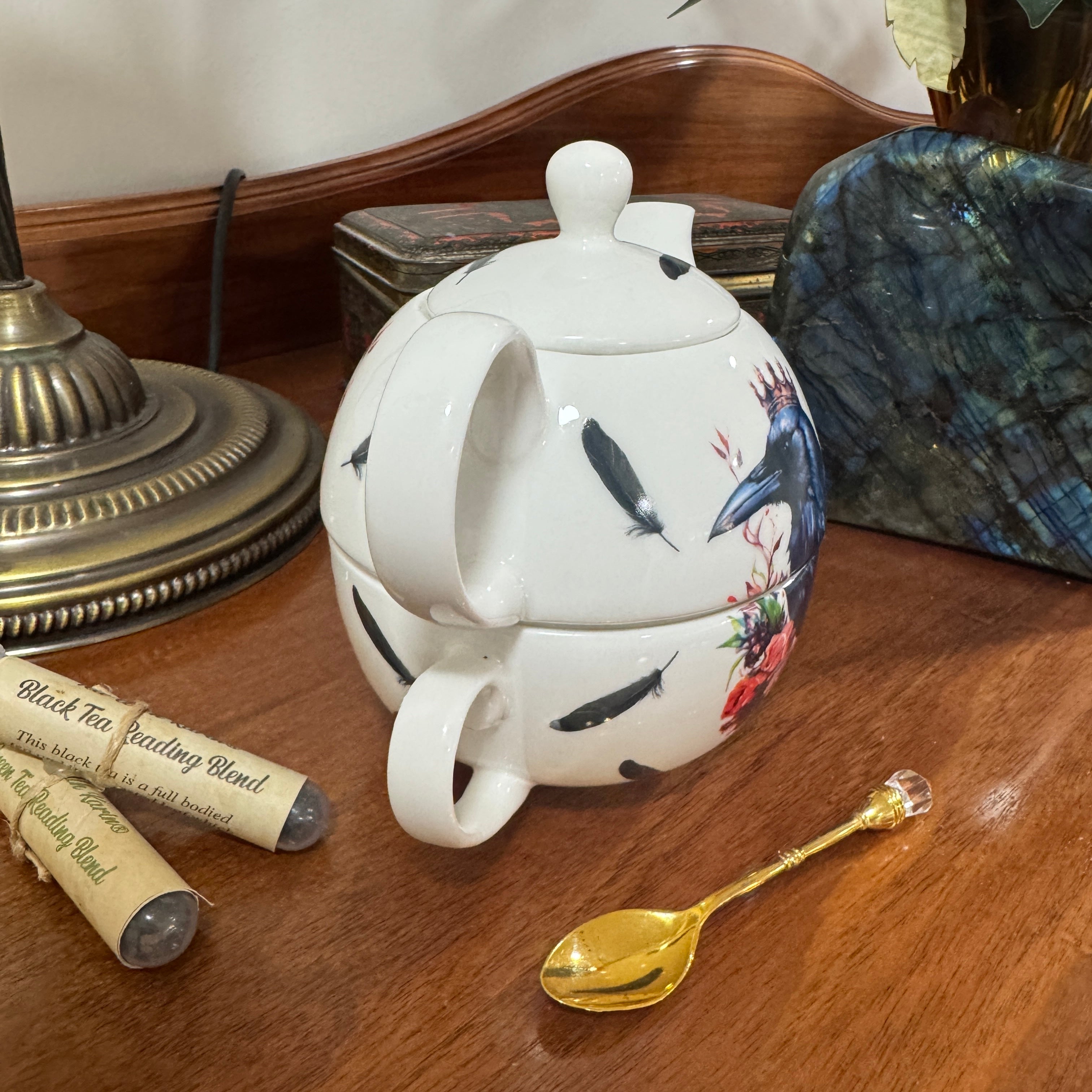 Decorative teapot with bird and floral design on a wooden surface with a gold spoon and tea packaging.