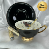Black and gold teacup and saucer with an internal skull-moth and classic tea-leaf reading symbols, a gold crystal spoon, and two tea sample tubes.