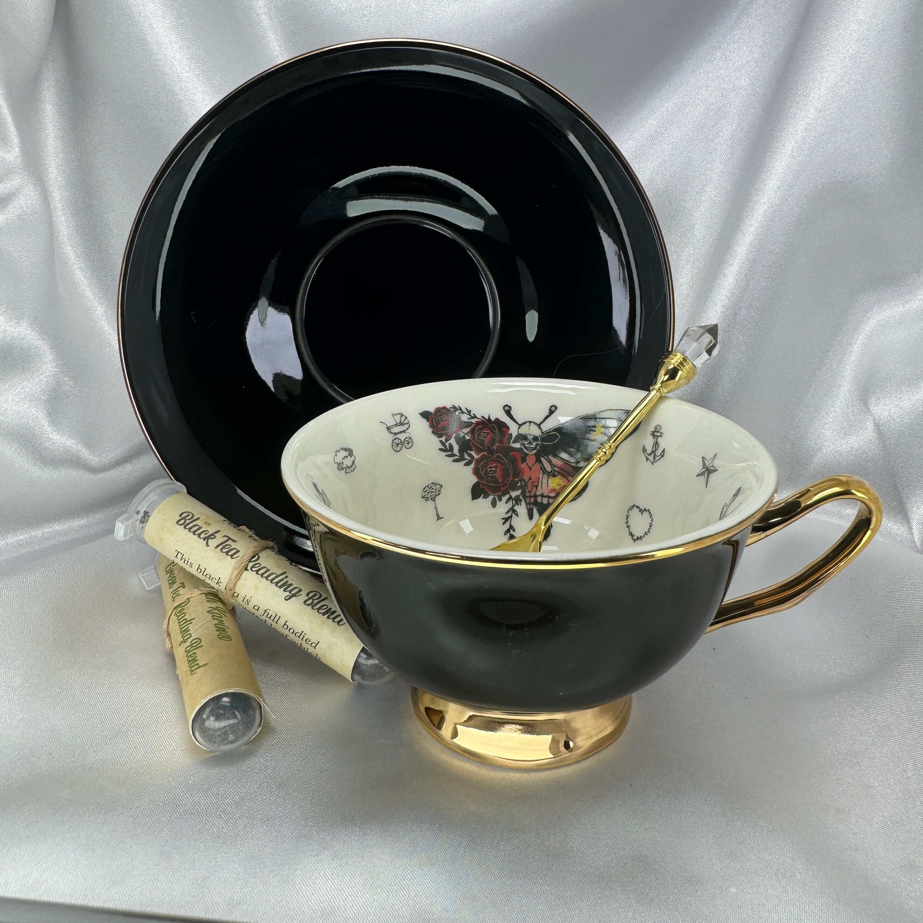 Black teacup with gold interior and handle, featuring a floral moth design, on a white fabric background.