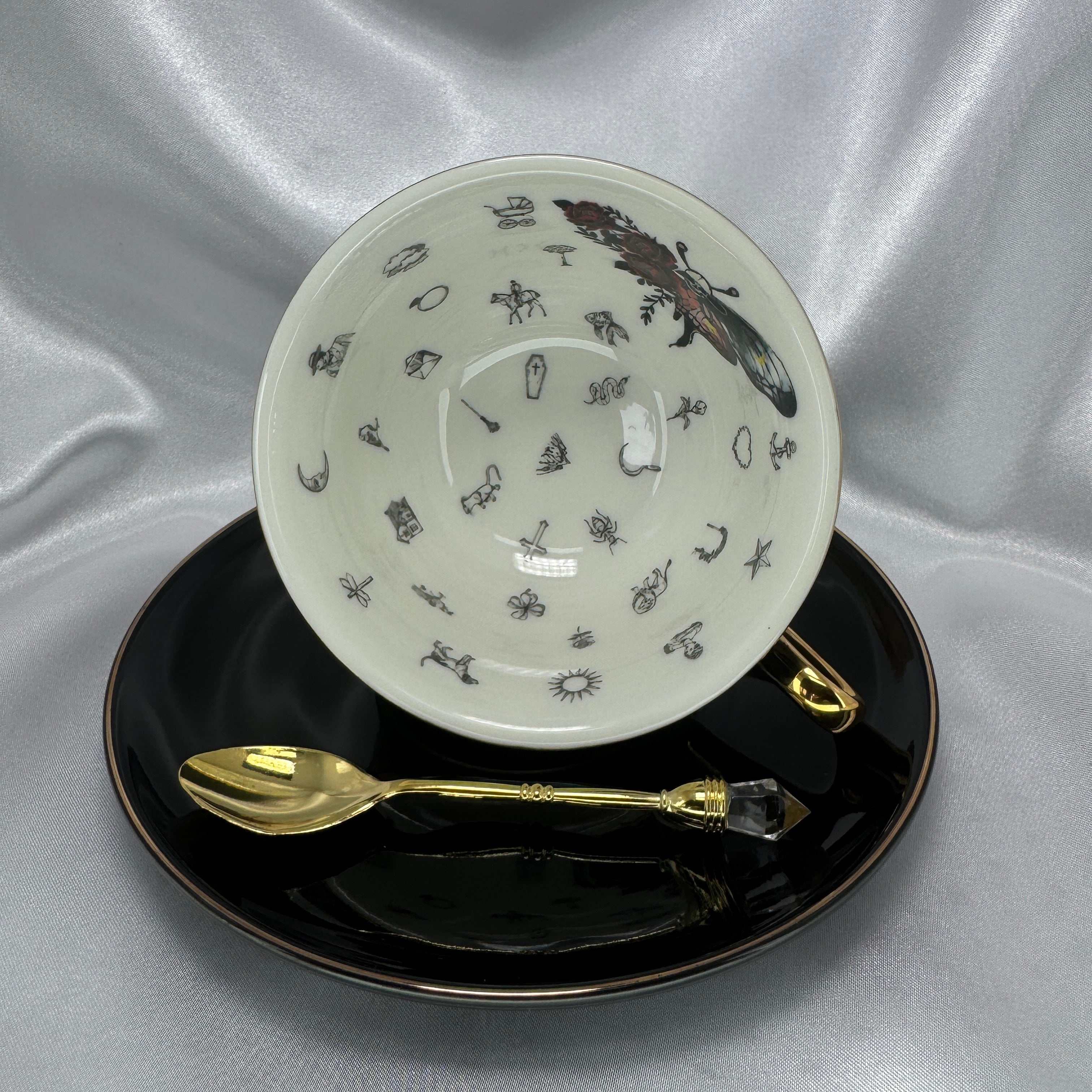 Teacup on its side showing the white interior with a large colourful skull-moth and small black classic fortune-telling symbols.