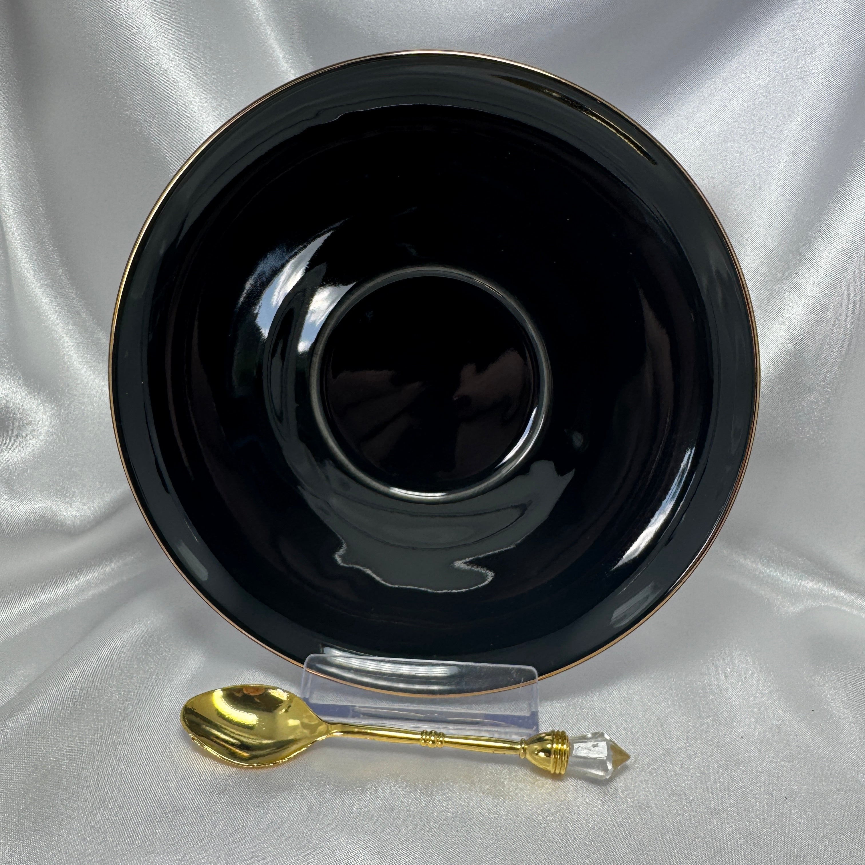 Shiny black saucer with a gold rim and a gold teaspoon with a clear crystal handle on a white satin background.