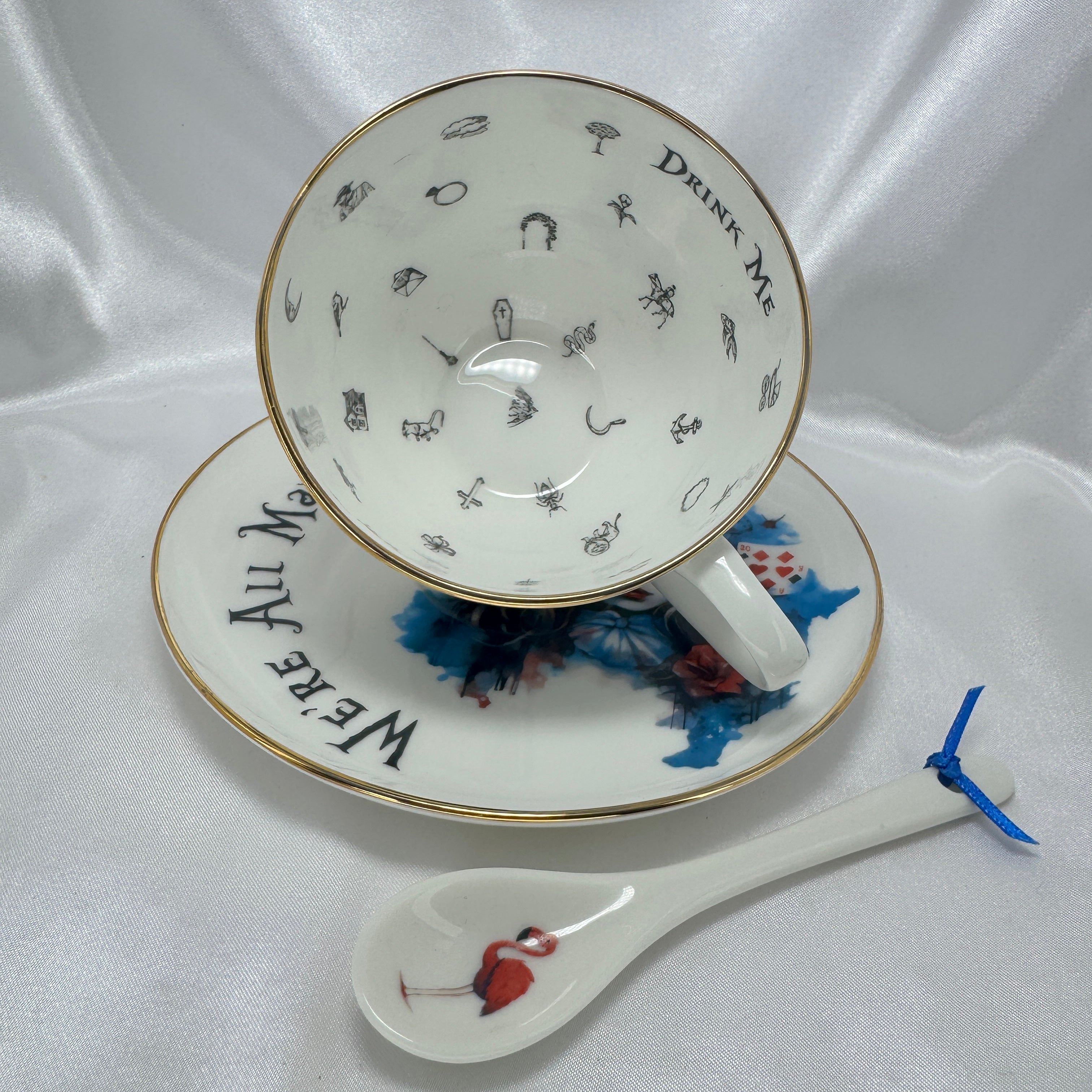 Tea cup and saucer with 'Drunk Me' and 'We're all mad here text, accompanied by a spoon with a flamingo design on a white fabric background.