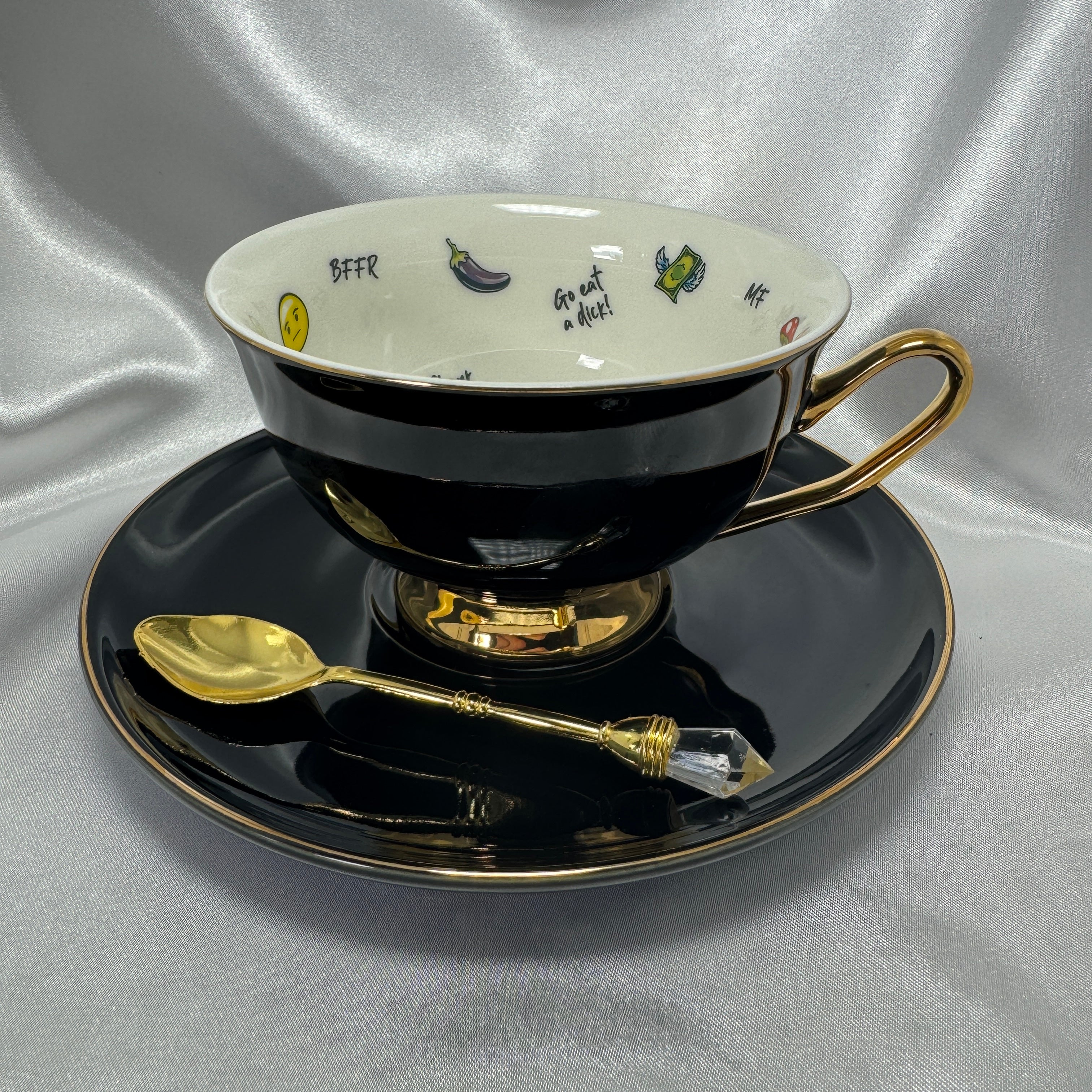 Teacup and saucer in black and gold with a gold teaspoon, showcasing the teacup's white interior and bold, humorous decoration.