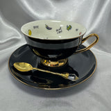 Teacup and saucer in black and gold with a gold teaspoon, showcasing the teacup's white interior and bold, humorous decoration.