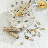 A styled flat-lay of a white porcelain teacup and saucer with gold trim and illustrated motifs, a gold spoon inside the cup, several silver and gold tea-themed charms, a rolled parchment label reading 'Black Tea Reading Blend,' and a gold circular badge that says 'Free Course' on a white wood-textured background.
