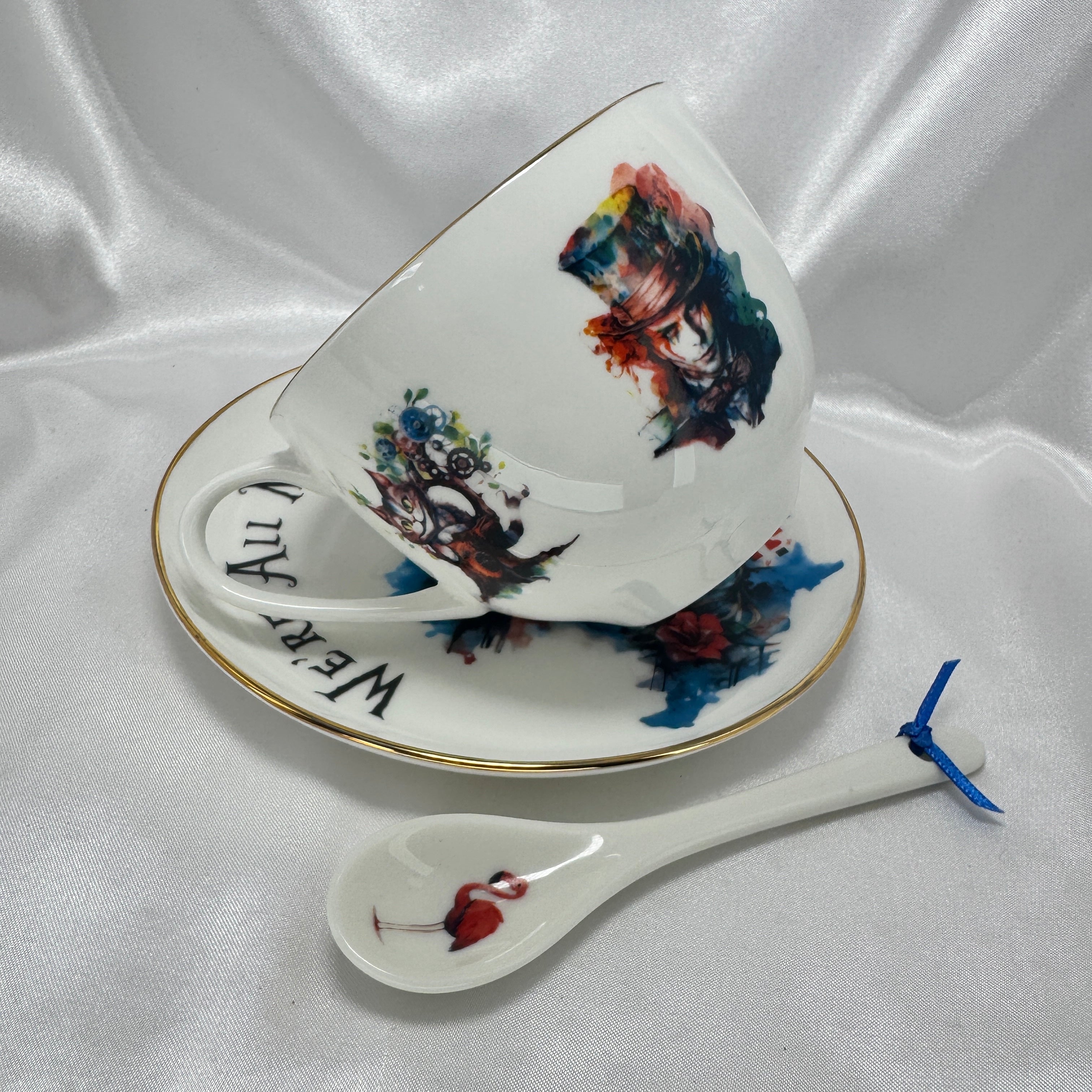Ceramic teacup, saucer, and spoon with artistic designs of Mad Hatter and Cheshire Cat on a white fabric background