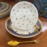 Decorative teacup with intricate designs on a saucer, accompanied by a gold spoon on a wooden surface.