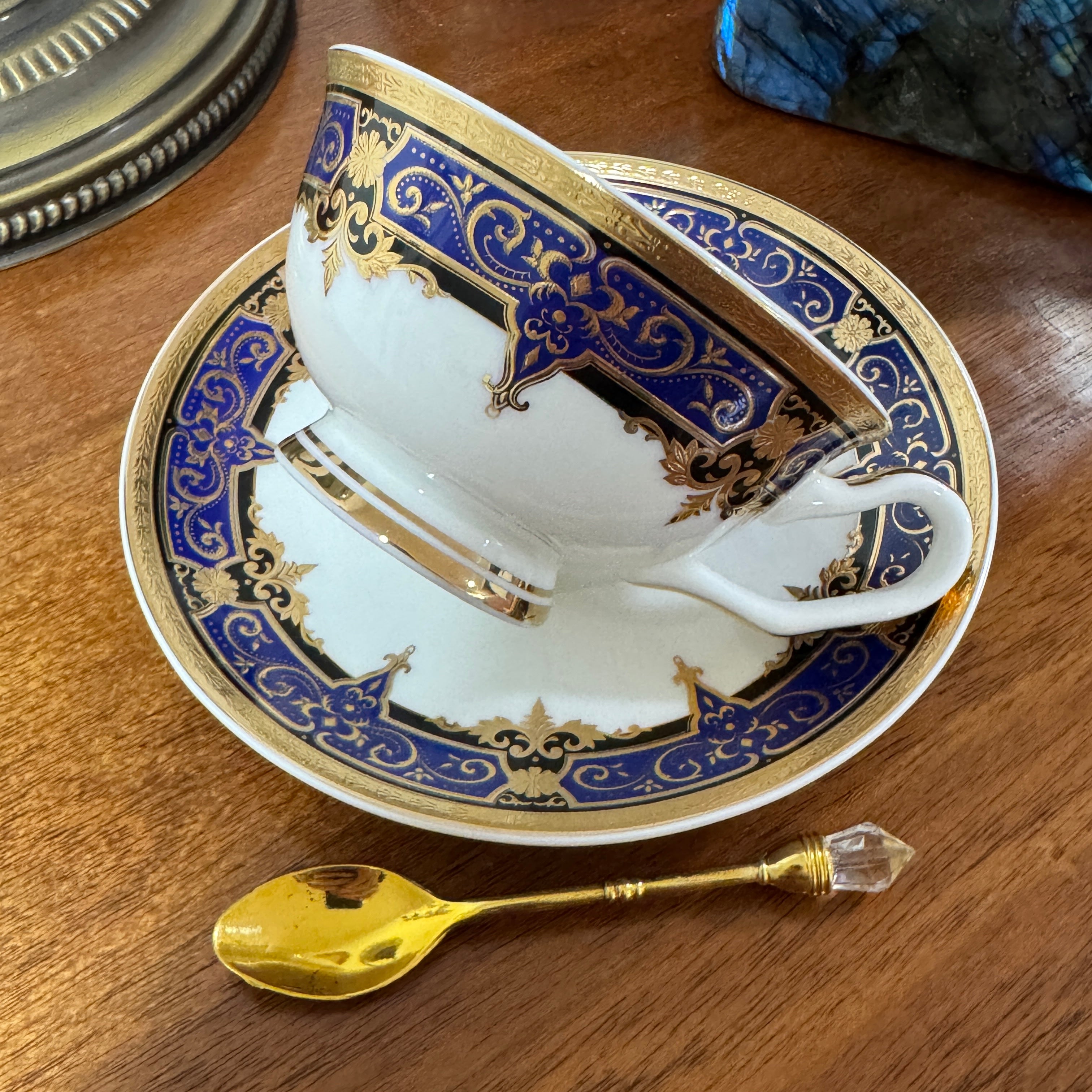 Decorative teacup and saucer with blue and gold patterns on a wooden surface, accompanied by a gold spoon.