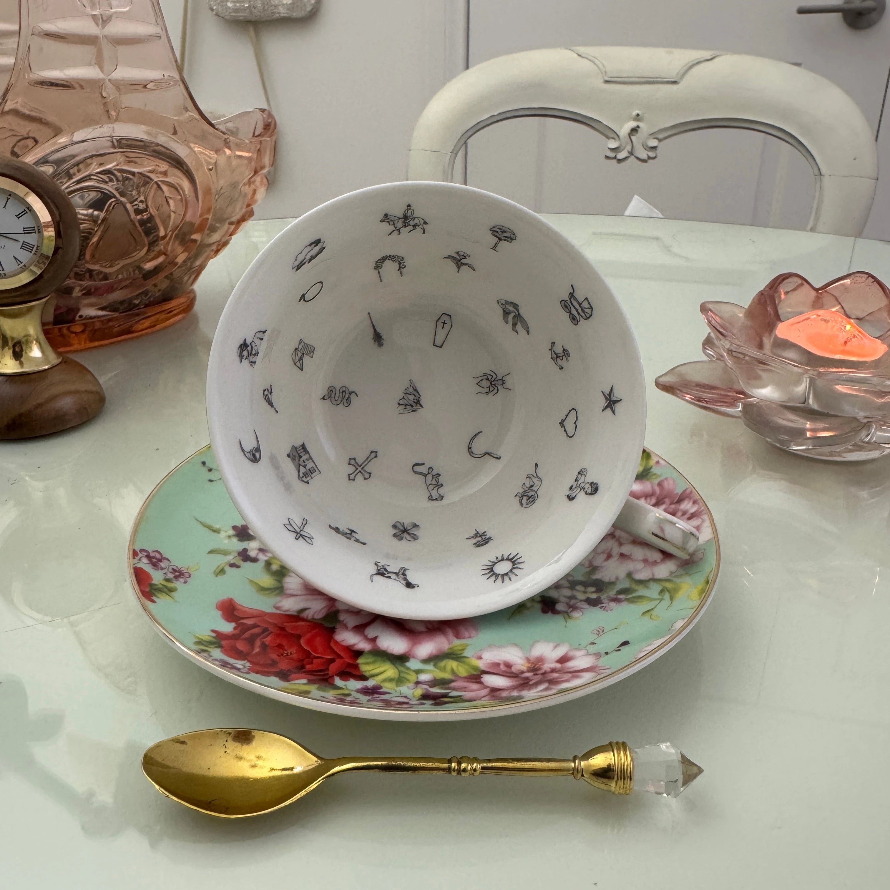 Inside view of mint red rose floral tasseography teacup face-up showing all 32 printed tea leaf reading divination symbols, resting on matching saucer with gold crystal teaspoon