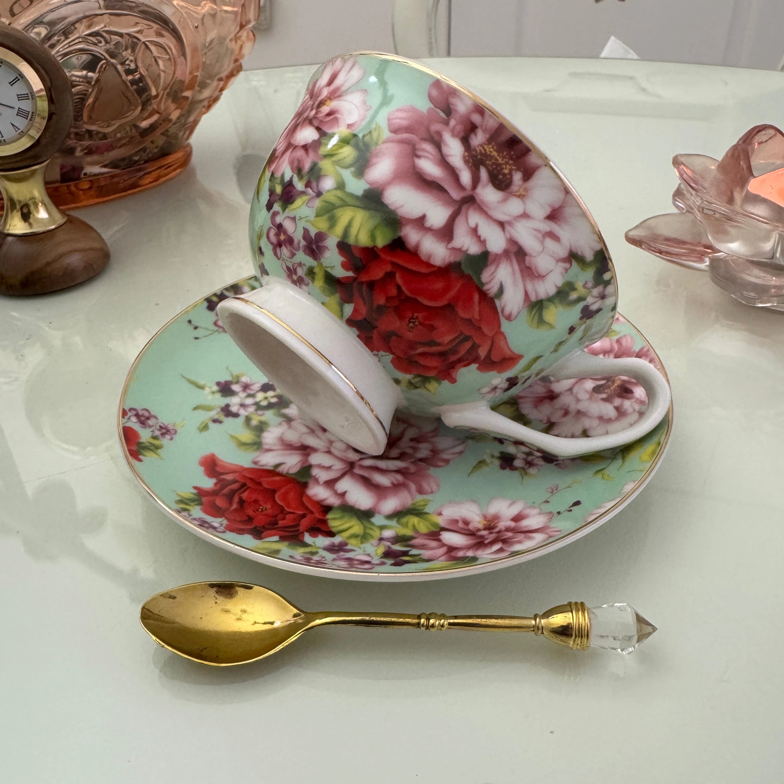 Left side view of mint red rose floral tasseography teacup tilted on matching saucer with gold crystal teaspoon beside it