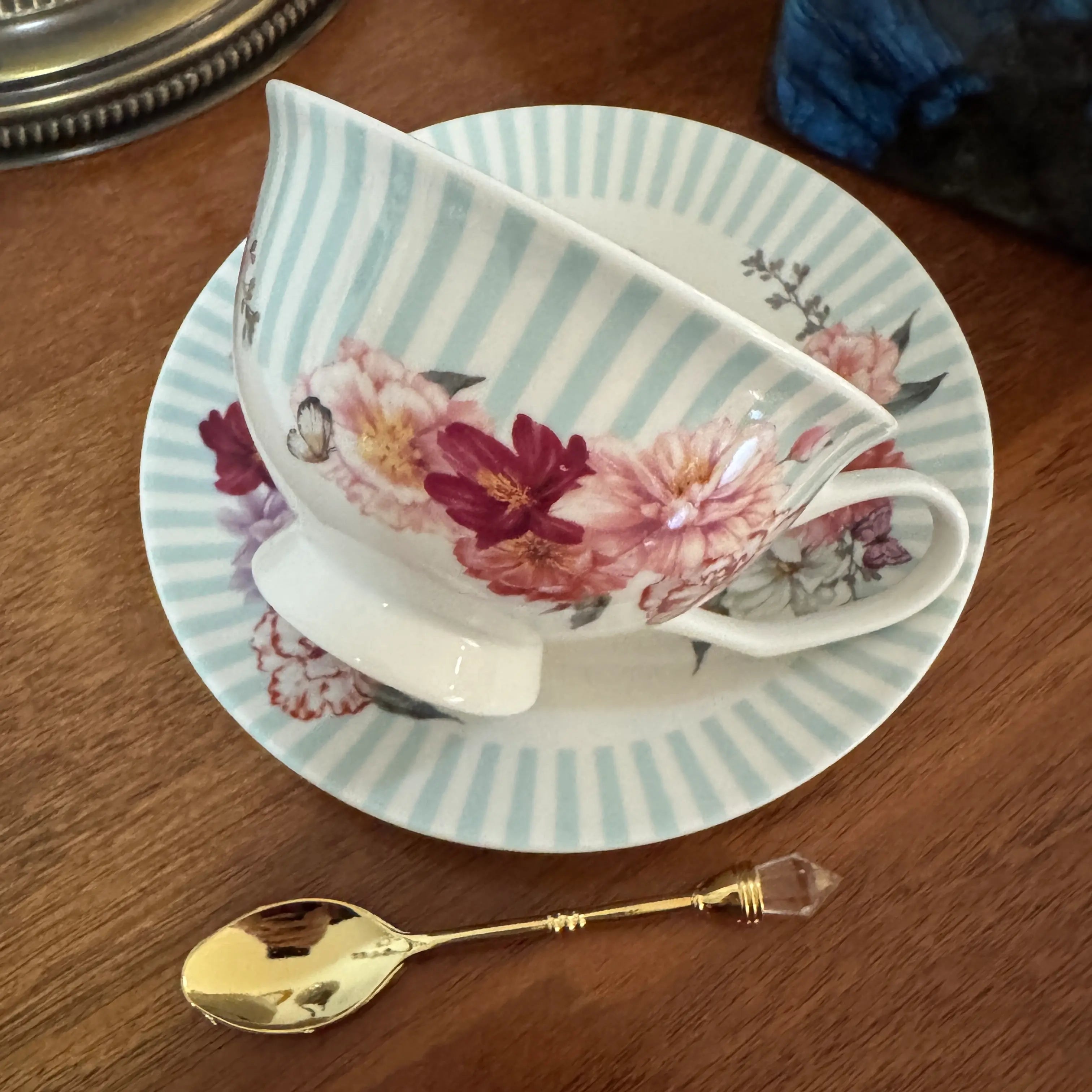 A white porcelain teacup and saucer set with light blue vertical stripes and pink floral patterns, displayed on a wooden table next to a gold spoon.