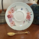 A white ceramic saucer plate with a light blue striped border, decorated with pink and red floral patterns and small butterflies, displayed on a wooden table next to a gold spoon.