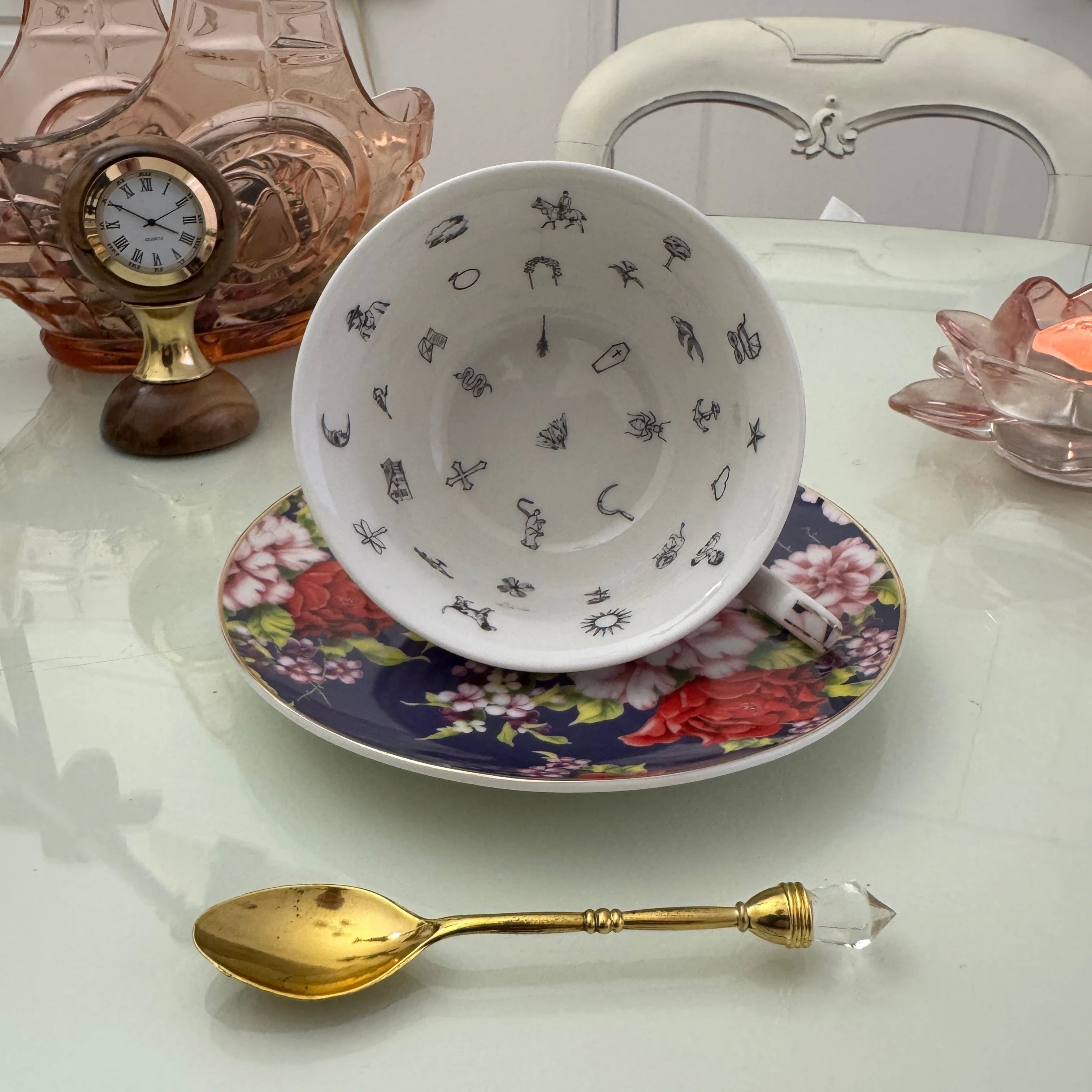Inside view of navy red rose floral tasseography teacup face-up showing all 32 printed tea leaf reading divination symbols, resting on matching saucer with gold crystal teaspoon