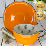 Bright orange teacup and saucer with silver rim and handle, decorative teaspoon, and two small tubes of Black Tea Reading Blend on a white wooden surface.