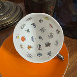 A white ceramic teacup decorated with small colorful tarot illustrations, shown resting on an orange saucer on a wooden table.