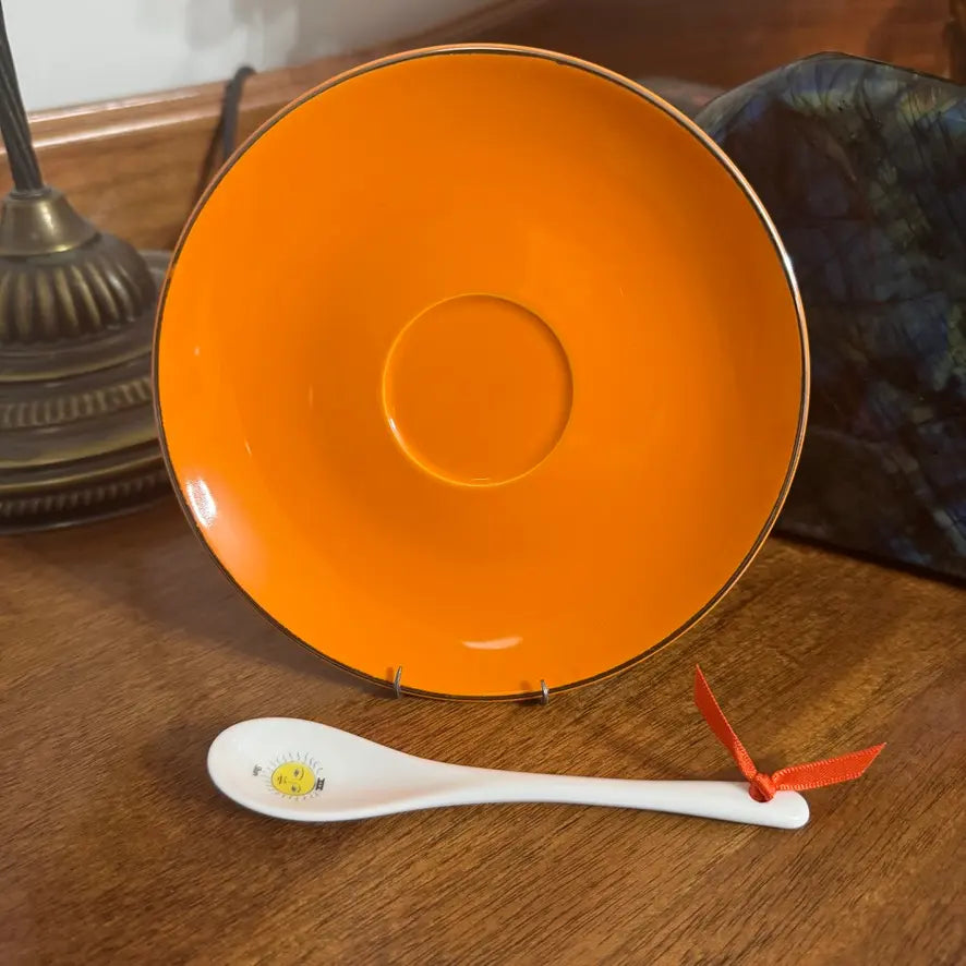 An orange ceramic bowl with a hand-painted silver rim displayed on a wooden surface, with a white ceramic spoon featuring a small yellow sun illustration and a red ribbon tied to its handle in the foreground.