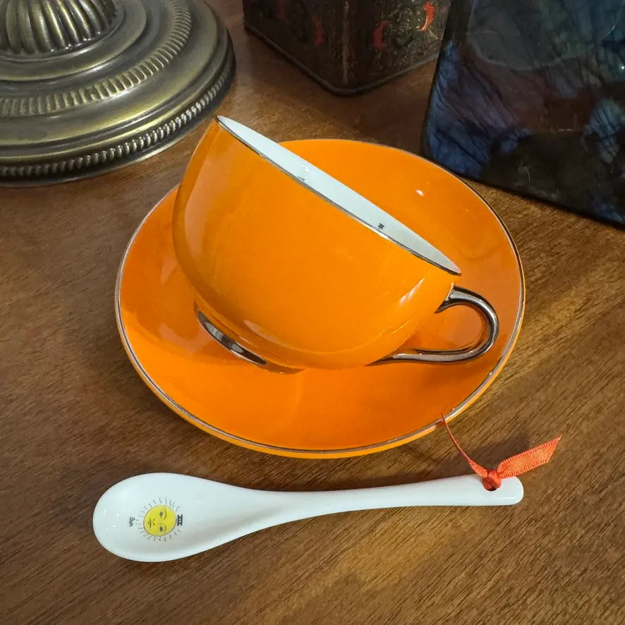 An orange ceramic teacup and saucer set with a white spoon featuring a small yellow smiley face, displayed on a wooden tabletop.