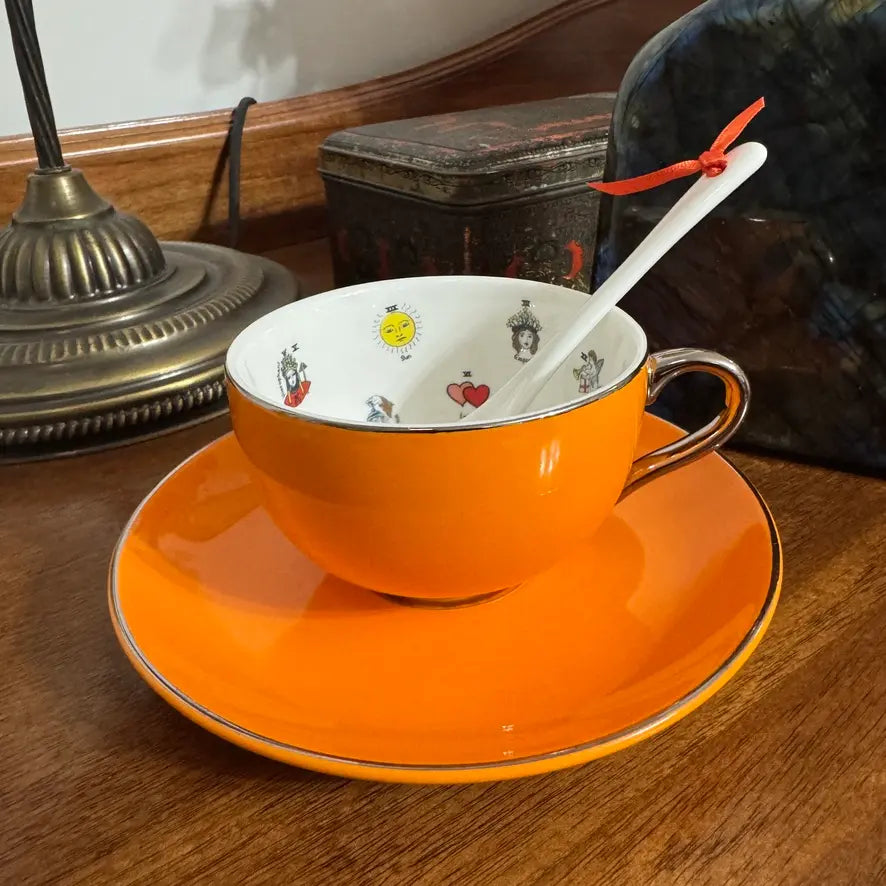 An orange ceramic teacup and saucer set with a white spoon, photographed on a wooden table. The cup and saucer are bright orange with a thin metallic rim, and the inside of the cup is white with small decorative illustrations.