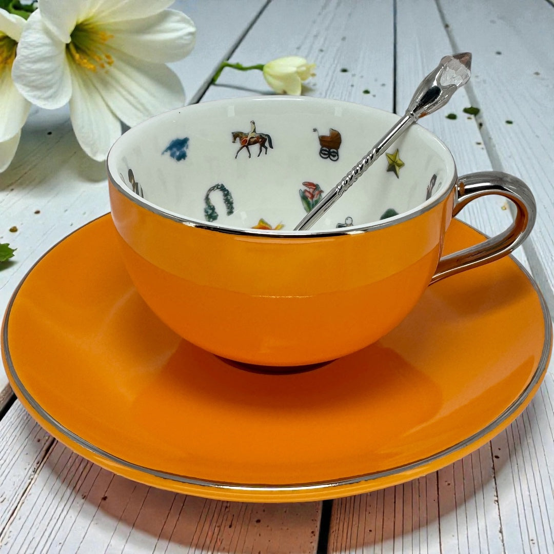 A vibrant orange tea cup and saucer set with a decorative silver spoon sitting on a white wooden table with white lilies.