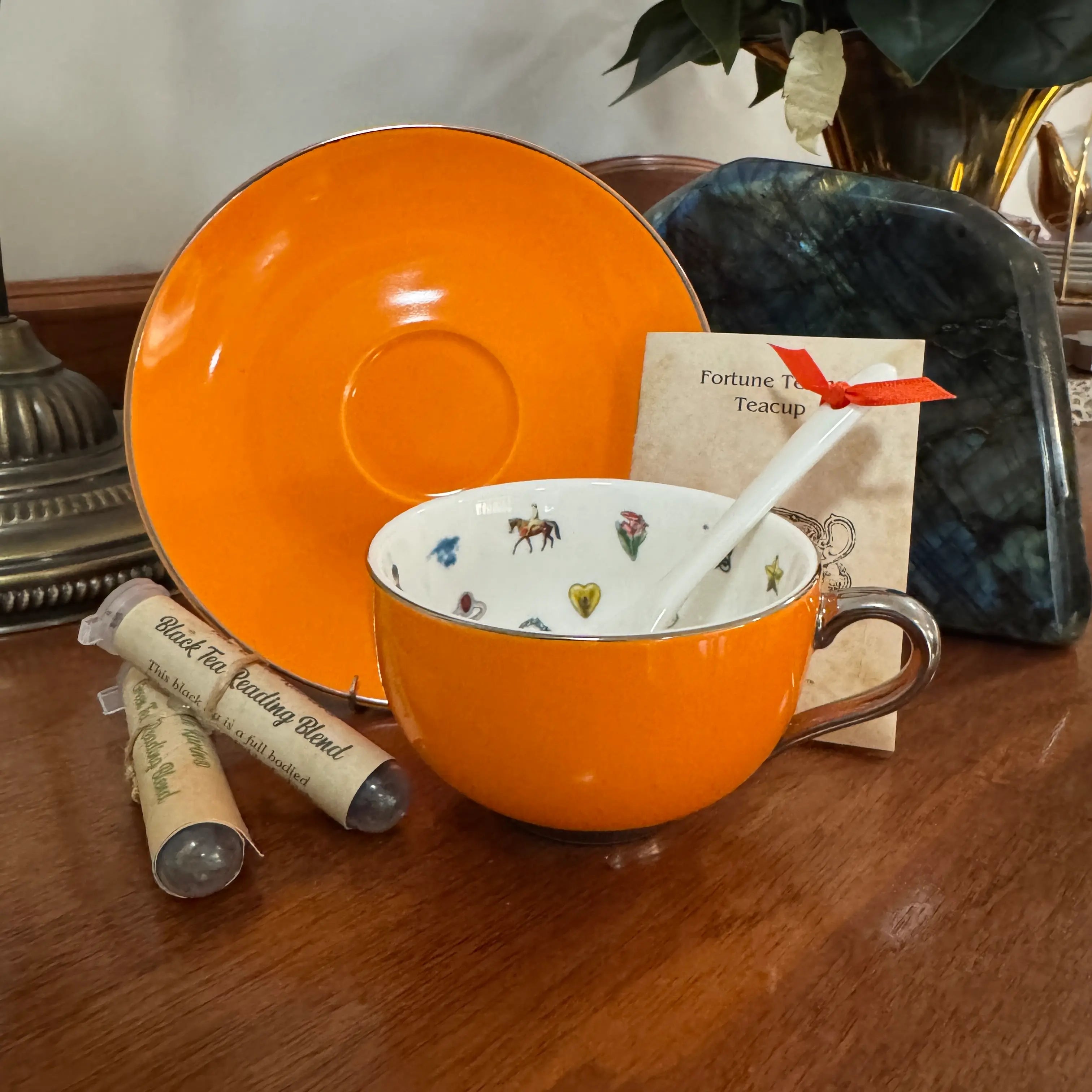 An orange ceramic teacup and saucer set on a wooden table, accompanied by a white spoon, a small card labeled Fortune Tea Teacup, and two wrapped tea blends.