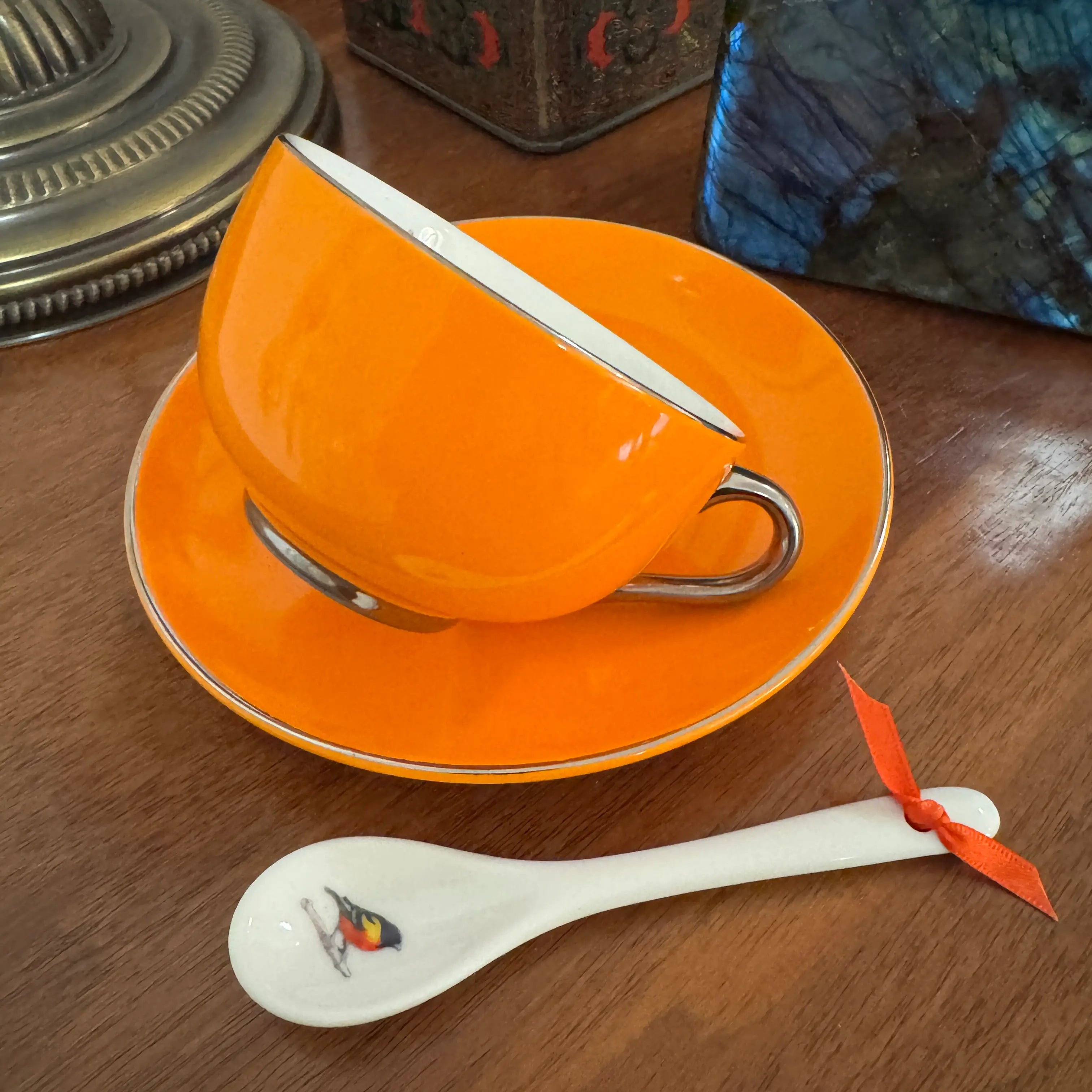 An orange ceramic cup and saucer set with a white decorative spoon on a wooden table. The cup and saucer have a glossy finish with a thin metallic rim, and the spoon is white with a small bird illustration and tied with an orange ribbon.