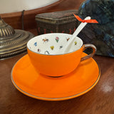 An orange ceramic teacup and saucer set on a wooden table, with a white spoon inside the cup and a small orange ribbon tied to the handle. The cup interior is white with small multicolored decorative motifs.