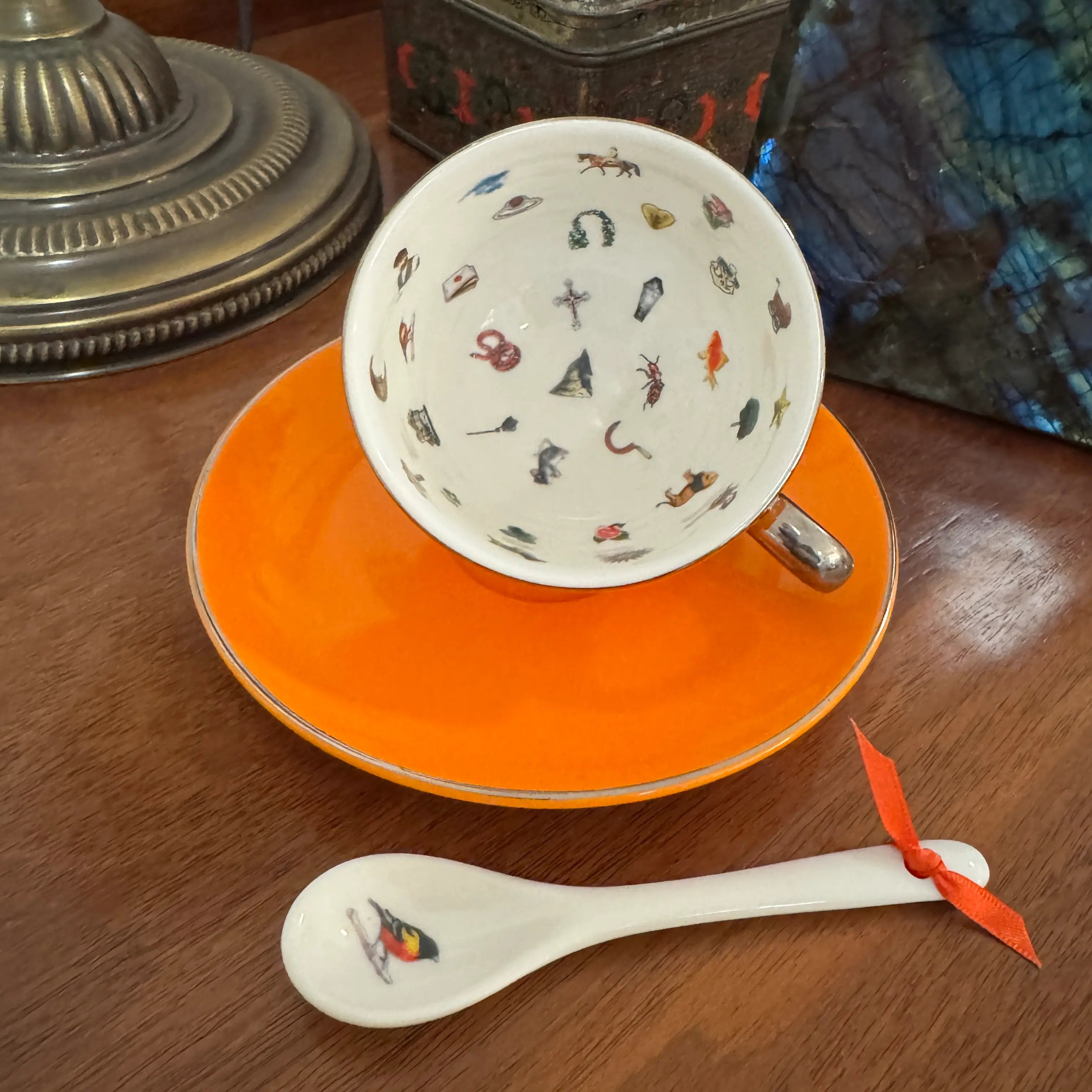 A white ceramic teacup and saucer set with a matching spoon, featuring a colorful pattern of small illustrated objects inside the cup, placed on a wooden table.