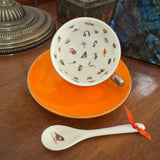 A white ceramic teacup and saucer set with a matching spoon, featuring a colorful pattern of small illustrated objects inside the cup, placed on a wooden table.