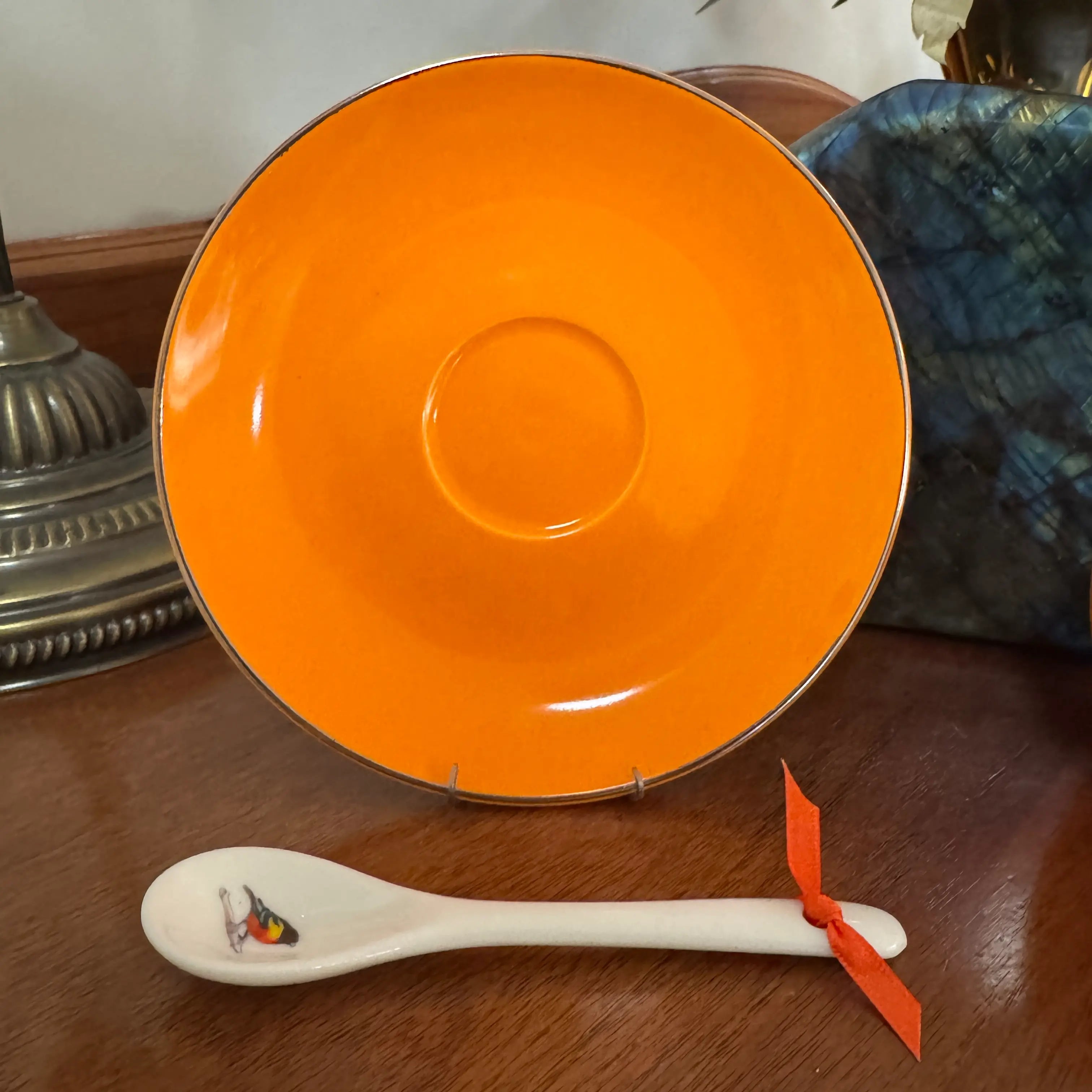 An orange ceramic plate with a silver rim displayed on a wooden table, with a white ceramic spoon featuring a small bee illustration and a red ribbon tied to its handle in the foreground.