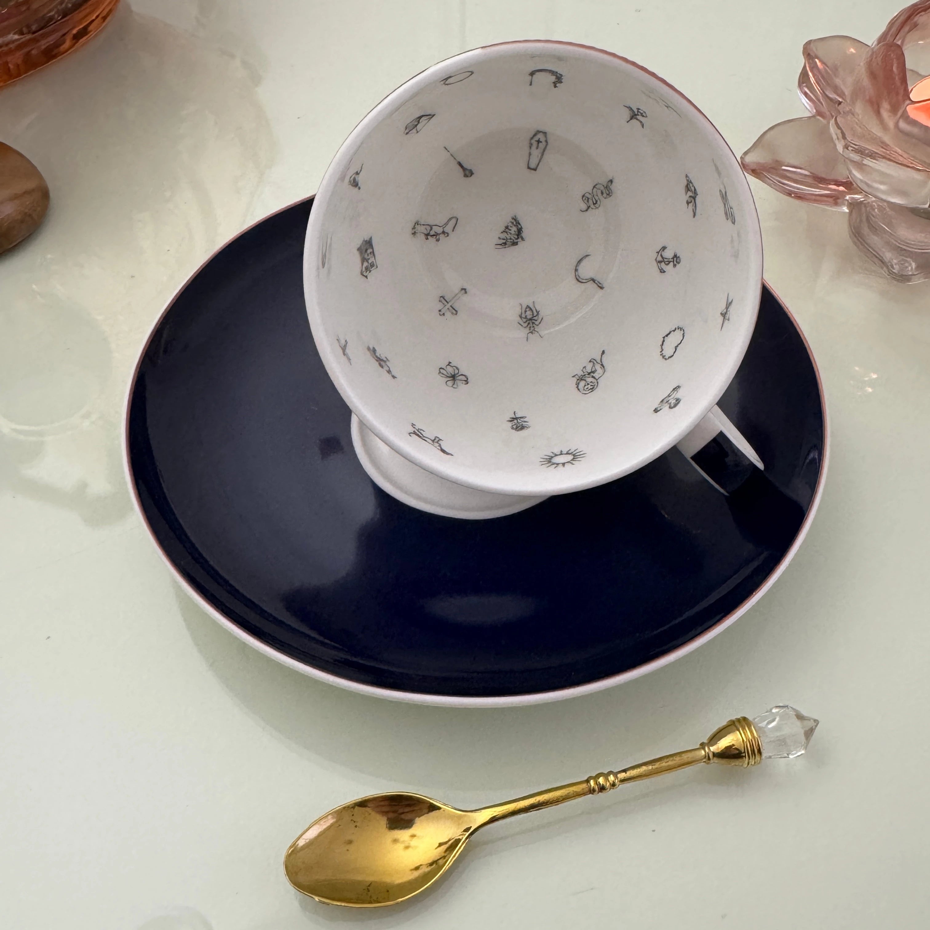 Inside view of peacock navy blue tasseography teacup face-up showing all 32 hand-painted tea leaf reading divination symbols, resting on matching saucer with gold crystal teaspoon
