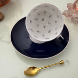 Inside view of peacock navy blue tasseography teacup face-up showing all 32 hand-painted tea leaf reading divination symbols, resting on matching saucer with gold crystal teaspoon
