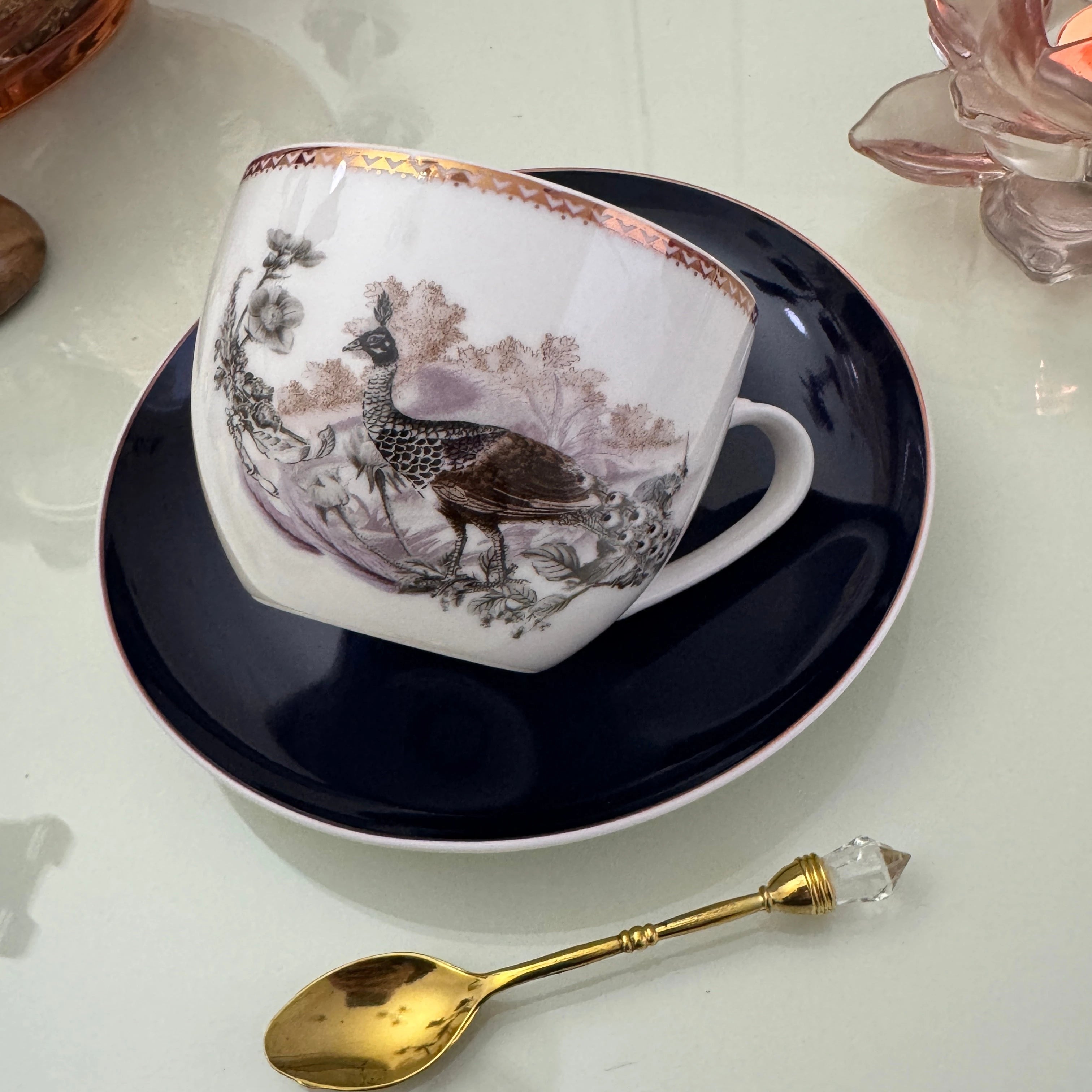 Left side view of peacock navy blue tasseography teacup tilted on matching saucer with gold crystal teaspoon beside it