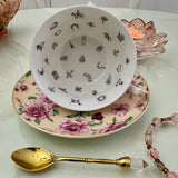 Inside view of pink butter rose floral tasseography teacup face-up showing all 32 printed tea leaf reading divination symbols, resting on matching saucer with gold crystal teaspoon