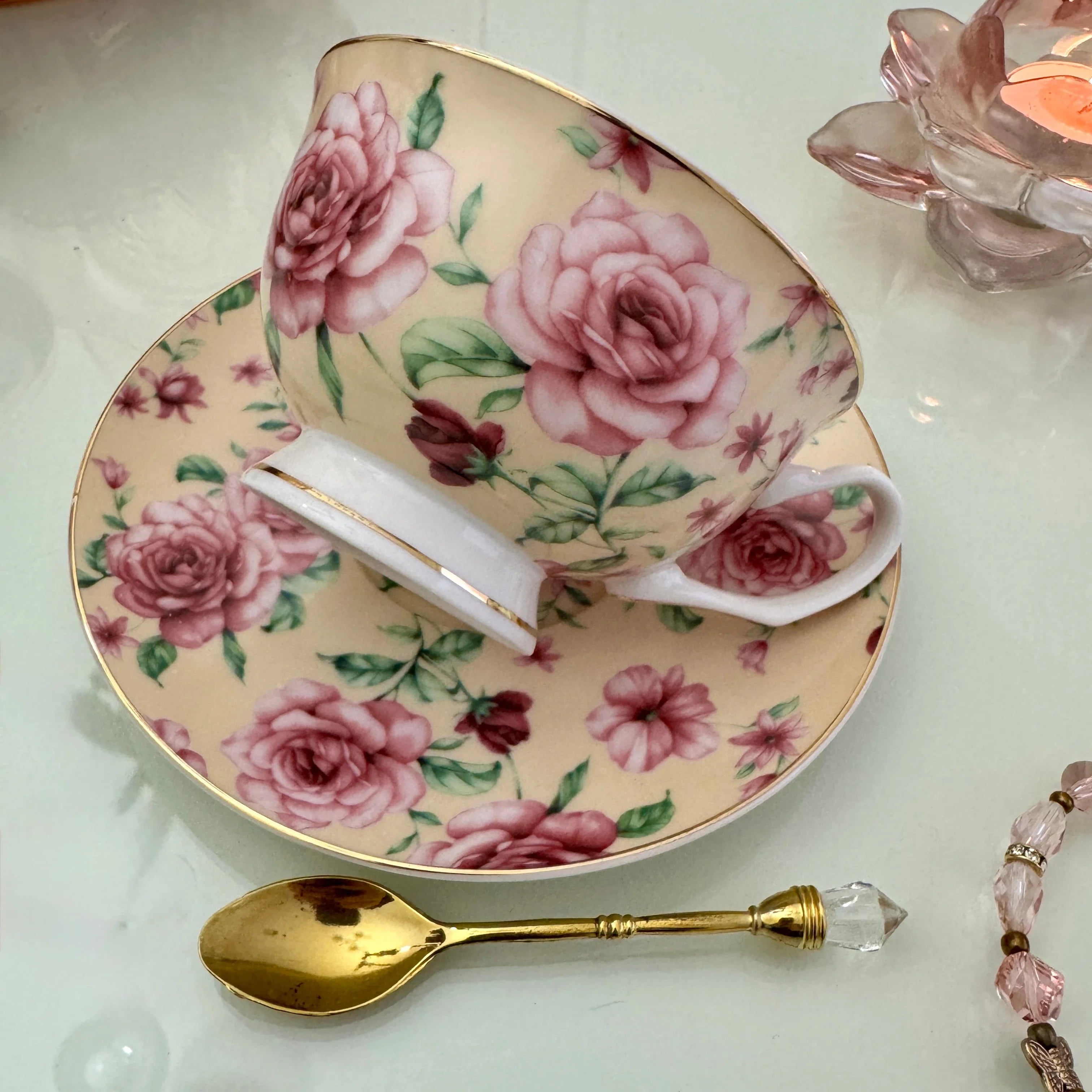 Left side view of pink butter rose floral tasseography teacup tilted on matching saucer with gold crystal teaspoon beside it